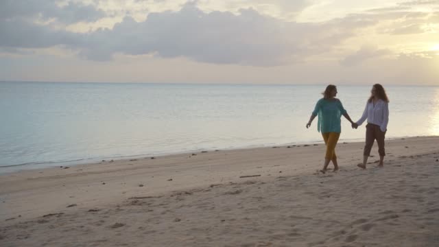 Zwei-junge-glückliche-Frauen,-die-bei-Sonnenuntergang-am-Strand-spazieren-gehen