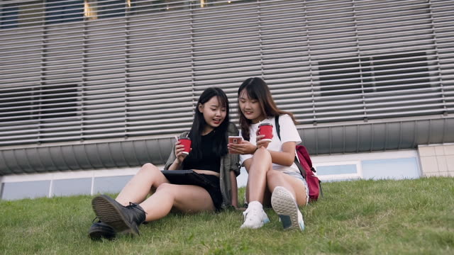 Frontansicht-der-charmanten-fröhlichen-asiatischen-Freundinnen,-die-auf-dem-grünen-Gras-sitzen-und-auf-das-Telefon-in-der-Nähe-von-modernen-Gebäude