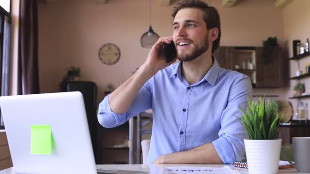 Sonriendo-hombre-de-negocios-feliz-usando-computadora-portátil,-sosteniendo-el-teléfono-inteligente,-hablando-en-el-teléfono-celular-se-sienta-en-el-escritorio,-haciendo-una-llamada-de-negocios-en-la-oficina-en-casa.