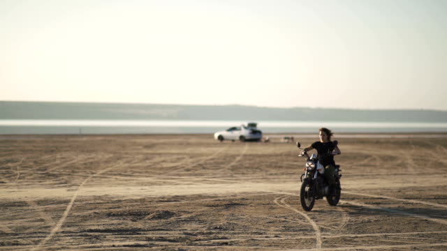 schöne-junge-Frau-mit-dem-Motorrad-fährt-auf-ein-altes-Motorrad-und-einen-Trick-zu-tun.-in-der-Wüste-bei-Sonnenauf--oder-Sonnenuntergang.-Weibliche-Biker.