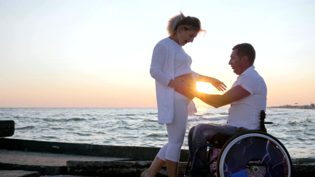 Pregnant-female-with-disabled-husband-in-wheelchair-at-sunset,-happy-disabled-person-caresses-woman-with-big-belly