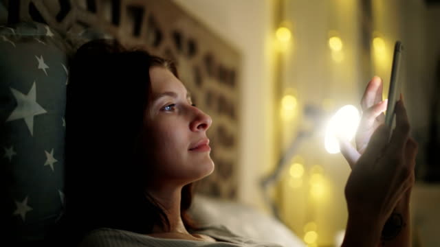 Lächelnde-Frau-spielen-online-Video-Spiel-auf-ihrem-Smartphone-nachts-am-Bett-zu-Hause-liegen