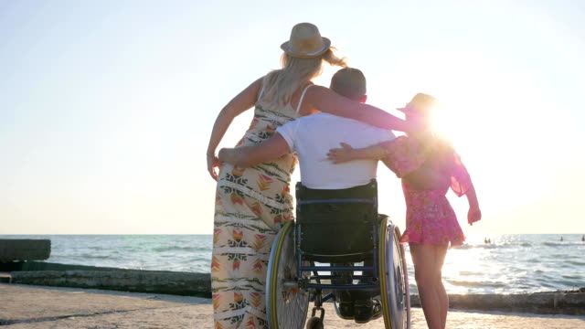 invalid-with-pregnant-woman-and-child-on-background-sea,-husband-in-wheelchair-hug-wife-with-big-belly