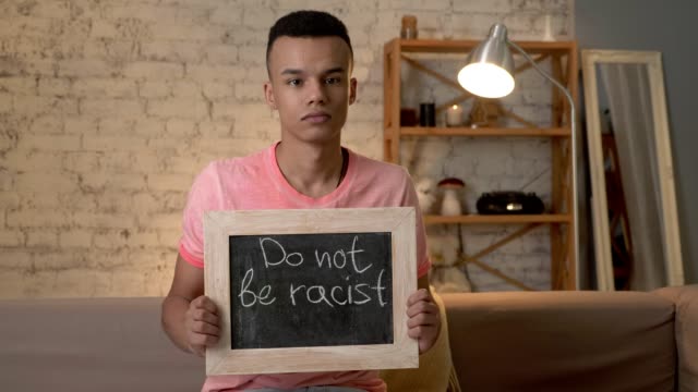 A-sad-African-guy-sits-on-the-couch-and-holds-a-sign.-Do-not-be-racist.-Anti-racism,-national-inequality-concept.-Look-at-the-camera.-60-fps