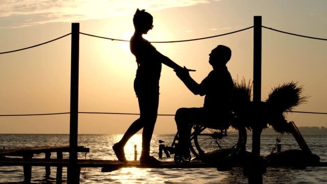 silueta-del-hombre-discapacitado-y-la-mujer-contra-el-sol,-el-romántico-encuentro-en-el-paseo-marítimo