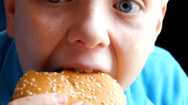 Un-muchacho-adolescente-come-una-hamburguesa-en-restaurante-de-comida-rápida,-junto-a-la-vista.-Boca-de-un-niño-que-come-una-hamburguesa