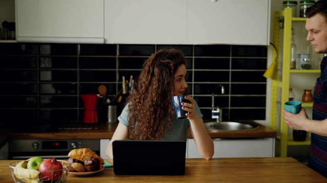 young-attractive-curly-happy-caucasian-woman-is-sitting-in-kitchen-working-on-laptop-drinking-tea,-her-husband-comes-and-she-shows-something-in-laptop