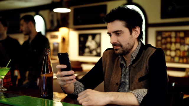 Young-male-student-is-using-smart-phone-sitting-in-fancy-bar-with-bottle-of-beer.-He-is-touching-screen-and-smiling.-Modern-ways-of-communication-concept.