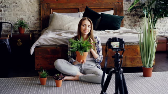 Young-smiling-blogger-in-casual-clothing-is-holding-flowers,-talking-and-recording-video-blog-for-online-vlog-about-house-plants-using-camera-on-tripod.