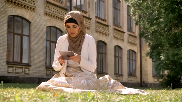 Beautiful-muslim-woman-in-hijab-typing-on-tablet-and-sitting-on-grass-in-park-near-building,-charming-female-with-gadget