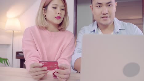 Young-Asian-family-couple-using-laptop-discussing-news-or-doing-online-shopping-sitting-together-on-desk-at-home-on-weekend-together,-serious-man-and-woman-browsing-web-searching-for-new-sale-offers.