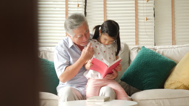 Asiatische-senior-Großvater-mit-einem-kleinen-Enkelin-Buch-zusammen-zu-Hause.-Familie-Senior-Lifestyle-Konzept.