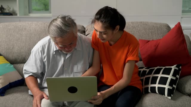 Daughter-teaching-her-father-computer-skills-in-living-room.-Asian-man-with-white-beard-and-young-woman-sitting-in-living-room-using-laptop.-Senior-lifestyle-family-concept.