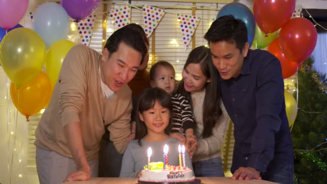 A-cute-little-girl-sitting-at-table-and-blowing-candles-on-birthday-cake-while-her-family-standing-behind-and-sing-a-song-to-her.-She-gives-a-big-smile-and-her-family-applaud-her.-In-slow-motion.