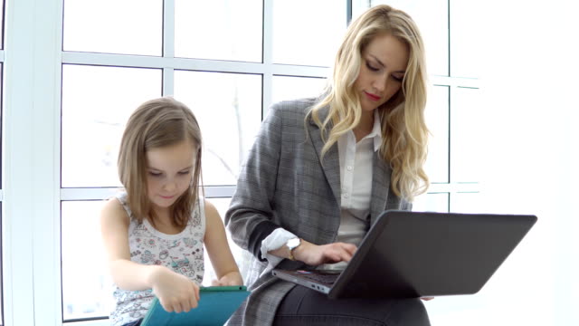 Junge-Geschäftsfrau-mit-Laptop-und-kleine-Mädchen-sitzen-am-Fenster.