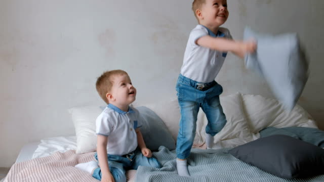 Twin-boys-toddlers-are-lying-on-the-bed,-throwing-pillows,-jumping-and-laughing.