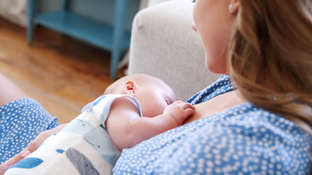 Close-up-shot-of-mother-sitting-on-sofa-at-home-breastfeeding-baby-son-viewed-over-her-shoulder---shot-in-slow-motion