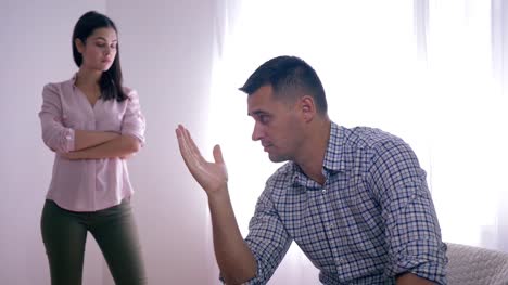 portrait-of-disappointed-guy-after-quarrel-with-female-sitting-at-home-folding-hands-near-face-and-woman-standing