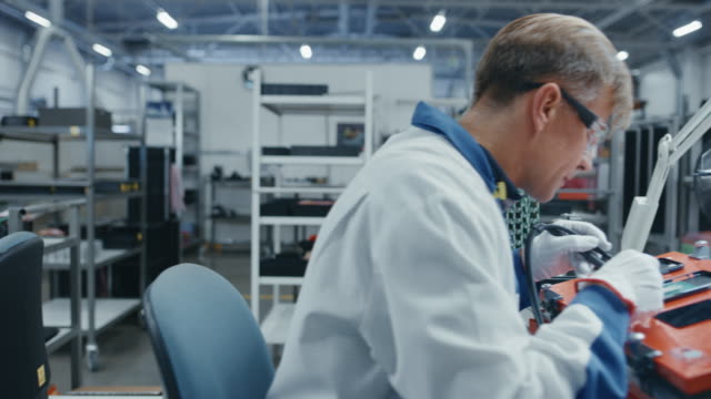 Time-Lapse-of-Young-and-Senior-Electronics-Factory-Workers-in-Blue-and-White-Work-Coats-Using-Pliers-and-Tweezers-to-Assemble-Printed-Circuit-Boards-for-Smartphones.-High-Tech-Factory-Facility.