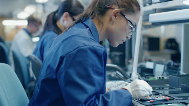 Weibliche-Elektronik-Fabrikarbeiter-in-blau-arbeiten-Mantel-und-Schutzbrille,-die-Montage-von-Leiterplatten-für-Smartphones-mit-einer-Pinzette.-Hightech-Fabrik-mit-mehr-Mitarbeiter-im-Hintergrund.