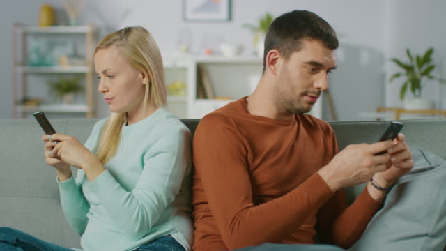 Entfremdung-/-streiten-Konzept:-zu-Hause-auf-ein-Sofa-junges-Paar-auseinander-sitzen,-mit-Blick-auf-unterschiedliche-Richtungen-und-Smartphone.