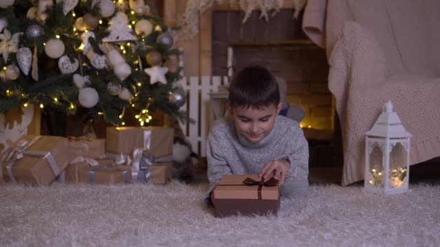 A-little-boy-opens-a-box-with-a-gift-and-rejoices-lying-on-the-floor-near-the-Christmas-tree