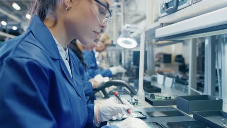 Woman-Electronics-Factory-Worker-in-Blue-Work-Coat-and-Protective-Glasses-is-Assembling-Smartphones-with-Tweezers-and-Screwdriver.-High-Tech-Factory-Facility-with-more-Employees-in-the-Background.