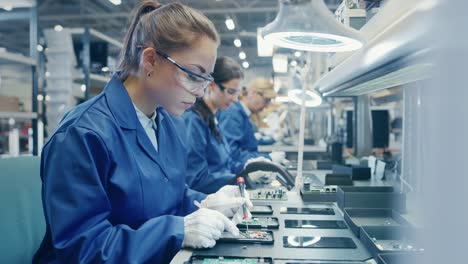Female-Electronics-Factory-Worker-in-Blue-Work-Coat-and-Protective-Glasses-is-Assembling-Smartphones-with-Tweezers-and-Screwdriver.-High-Tech-Factory-Facility-with-more-Employees-in-the-Background.