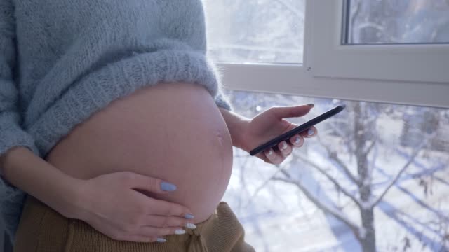 Schwangere-Mädchen-nutzt-Handy-und-streicheln-nackter-Bauch-im-stehen-durch-ein-großes-Fenster-in-Wohnung