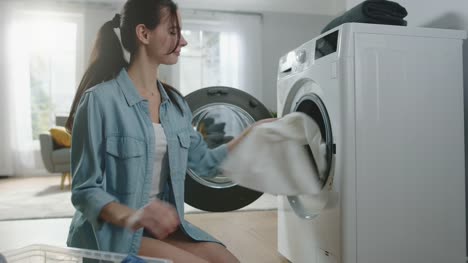 Beautiful-Smiling-Brunette-Young-Woman-Sits-in-Front-of-a-Washing-Machine-in-Homely-Jeans-Clothes.-She-Loads-the-Washer-with-Dirty-Laundry.-Bright-and-Spacious-Living-Room-with-Modern-Interior.