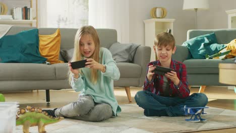 Zu-Hause-auf-einem-Teppich-sitzen:-niedliche-kleine-Mädchen-und-süße-junge-Videospiel-in-wettbewerbsfähige-auf-zwei-Smartphones,-hielt-sie-im-horizontalen-Modus-\"Querformat\".-Mädchen-gewinnt-und-feiert.
