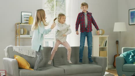 Two-Cute-Little-Girls-and-Young-Adorable-Boy-Have-Fun,-Jumping-High-on-a-Couch-at-Home.-Happy-Children-Playing-in-the-Sunny-Living-Room.-In-Slow-Motion.