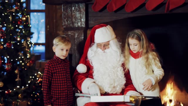 Santa-Claus-in-a-traditional-red-suit-sitting-in-the-middle-of-the-room-and-telling-stories-for-the-boy-and-girl-with-the-images-in-the-album,-which-he-is-holding-in-his-hands-near-the-fireplace-on-a-cold-winter-evening.
