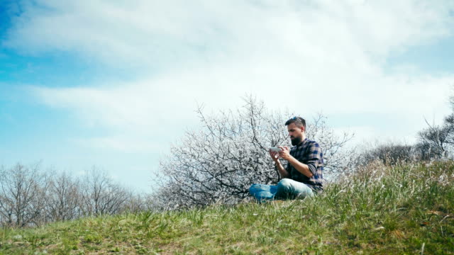 Hombre-con-una-barba-utiliza-una-tableta-en-naturaleza-cerca-de-flor-árbol