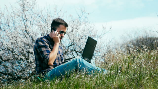 Dolly-shot.-Man-with-a-beard-uses-laptop-on-nature-near-blossoming-tree