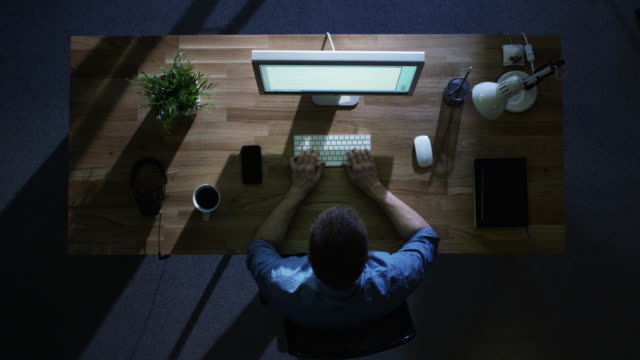 Vista-superior-del-programador-hombre-trabajando-en-su-computadora-de-escritorio-en-la-noche.-Su-mesa-está-iluminada-por-frío-azul-luz-desde-fuera.