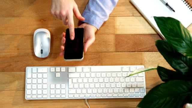 Hand-of-businesswoman-using-mobile-phone-at-desk-with-keyboard-and-mouse-on-table