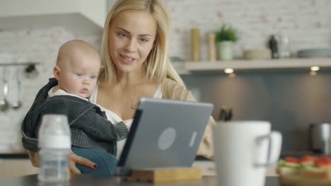 On-the-Kitchen-Mother-Holds-Her-Baby.-Both-Look-at-Something-Interesting-on-the-Screen-of-a-Tablet-Computer.