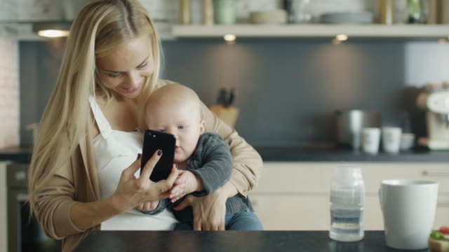 La-joven-madre-sostiene-a-su-bebé-mientras-está-en-la-cocina.-Ella-también-utiliza-smartphone-con-el-que-cute-Baby-interactúa.