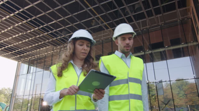 Team-von-zwei-Ingenieuren,-die-laufen,-reden-und-Tablet-Computer-nutzen.-Glasbau-oder-Wolkenkratzer-unter-Bau-auf-dem-Hintergrund.