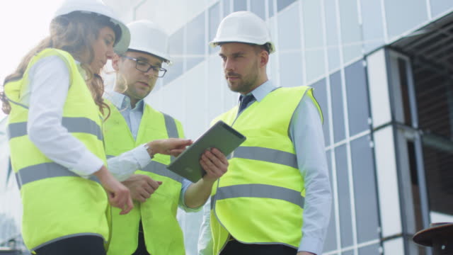 Team-of-Three-Engineers-having-Conversation-and-Using-Tablet-Computer.-Glass-Building-under-Construction-on-Background.