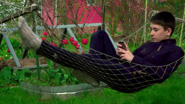 4K-Portrait-boy-Playing-Tablet-in-Hammock,-the-young-man-Face-Use-Smart-Phone-in-Nature