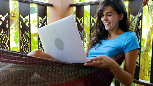 Mujer-joven-con-ordenador-portátil-mientras-se-relaja-en-la-hamaca-en-la-terraza-de-verano-escribiendo-feliz-sonriente,-chica-morena,-navegación-en-línea