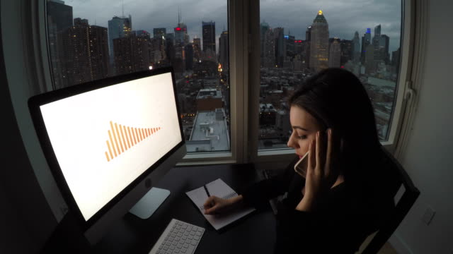 Attractive-Brunette-Working-at-Office-Table-at-Night.-Businesswoman-Working-with-Computer-and-Smartphone-in-Office-with-Cityscape-View.