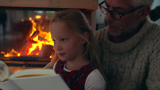Little-girl-reading-book-with-grandfather-near-fireplace