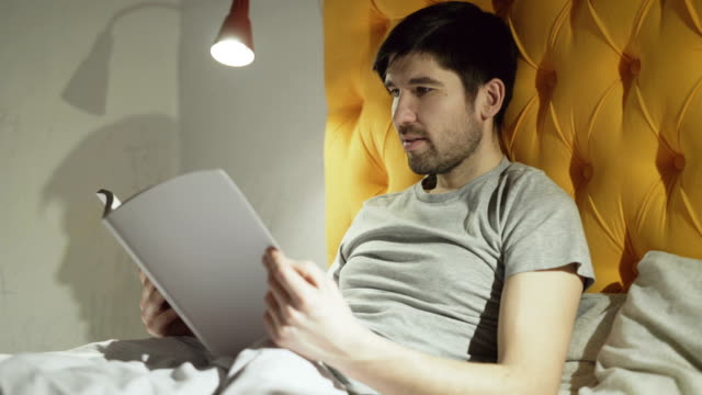 Young-father-reading-book-while-his-little-son-jumping-on-bed-before-sleeping-in-evening