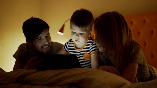 Familia-feliz-con-pequeño-hijo-aprendiendo-a-tocar-la-tablet-PC-tumbado-en-la-cama-en-casa-de-noche