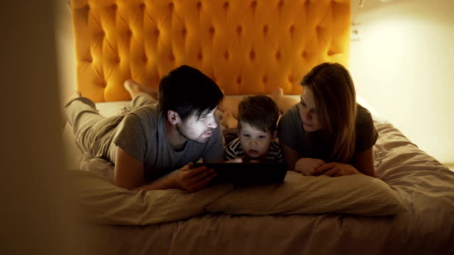 Glückliche-Familie-mit-kleinen-Sohn-eine-liegend-im-Bett-zu-Hause-und-mit-Tablet-PC-für-Cartoon-Film-vor-dem-schlafen