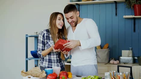 Cheerful-woman-surprising-his-boylfriend-with-birthday-gift-at-home-in-the-kitchen-while-he-cooking-breakfast