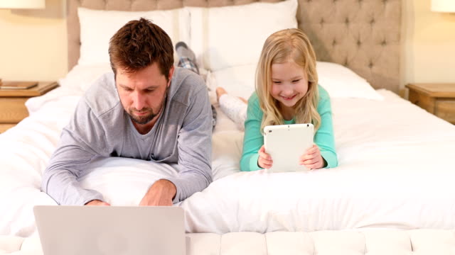 Glücklicher-Vater-und-Tochter-am-Bett-mit-Laptop-und-tablet-4K-4k